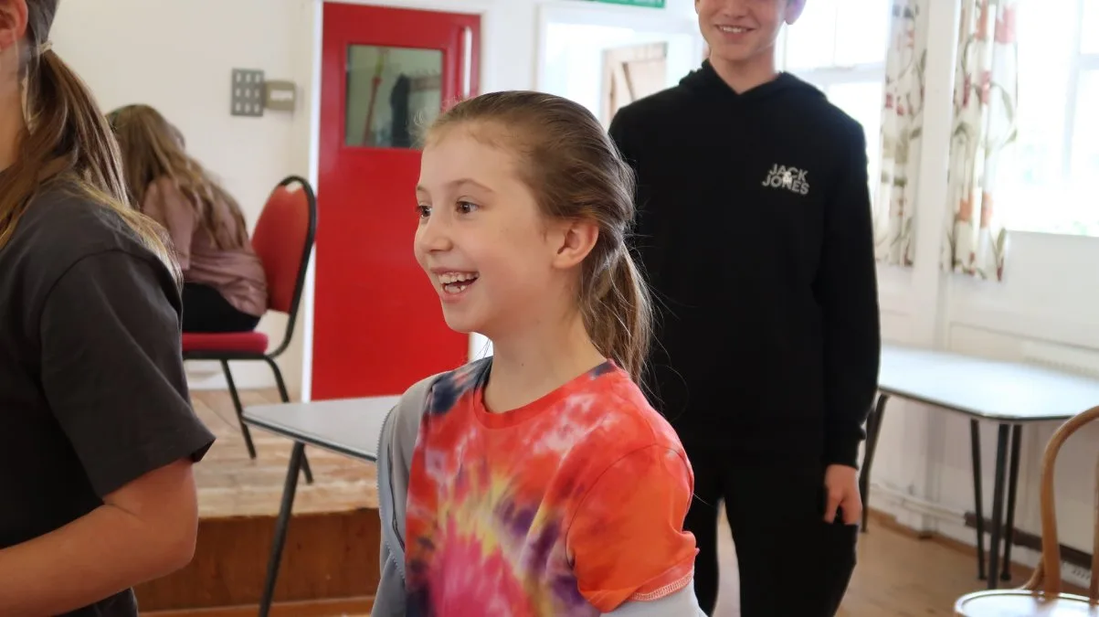 A smiling girl in a tie-dye top in a hall during a Sussex Youth Theatre session, with other young people around her