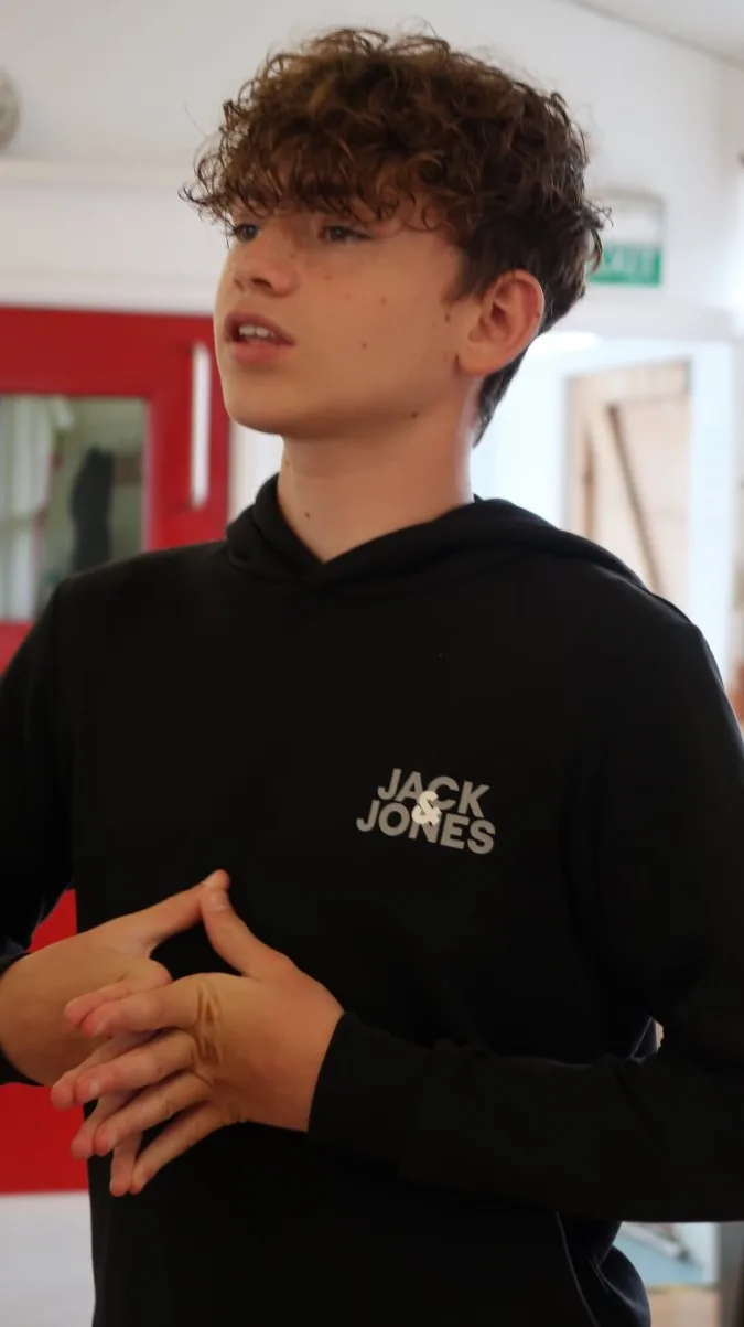 A teenage boy with curly hair in a black hoodie speaking during a drama session, hands clasped in front of him